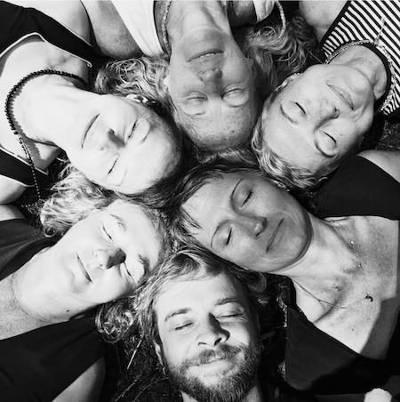 Six people lying in a circle with heads together, eyes closed, smiling, seen from above in black and white.