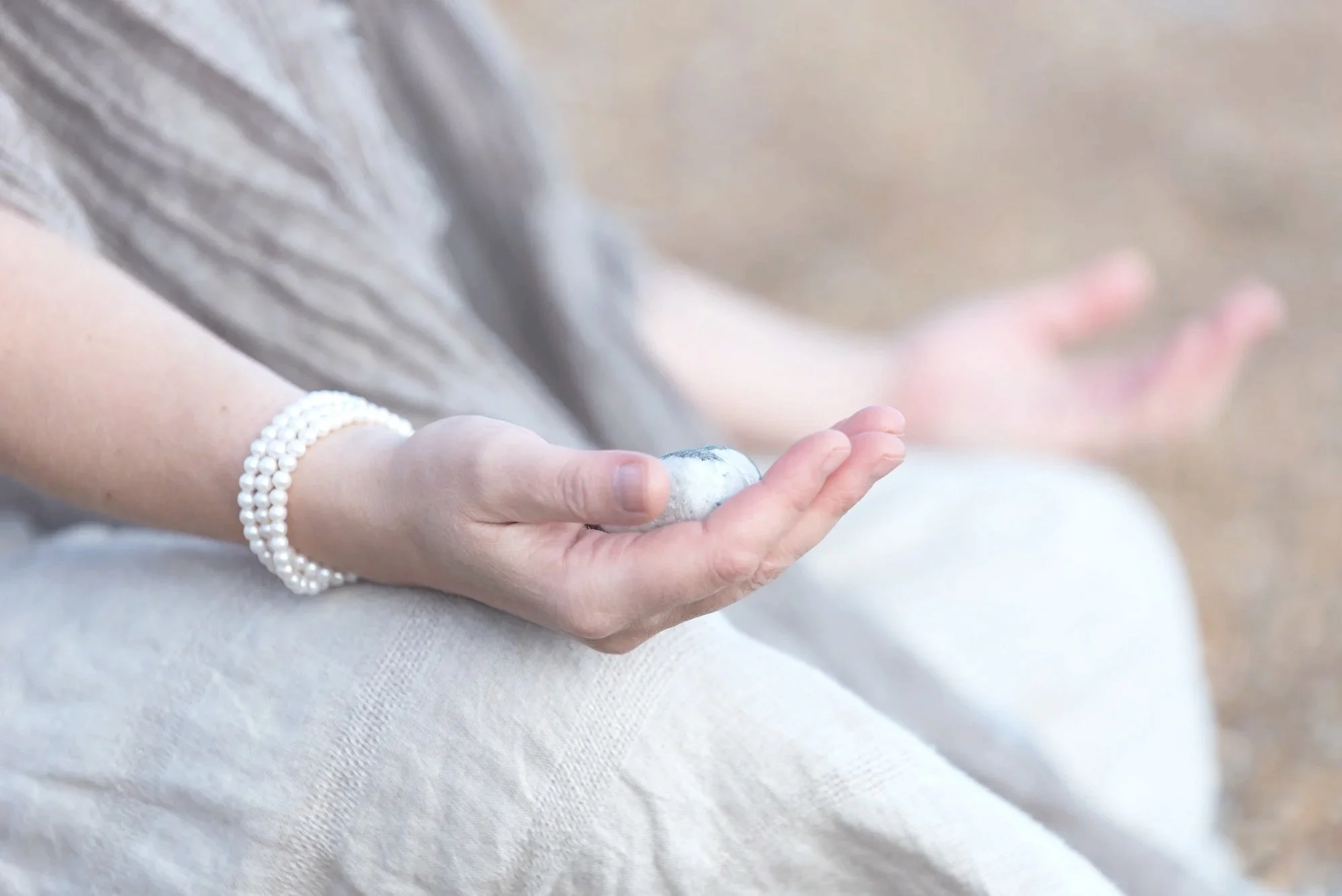 Close-up of a woman's hand resting on a light-colored fabric, wearing a pearl bracelet, with a blurred background.