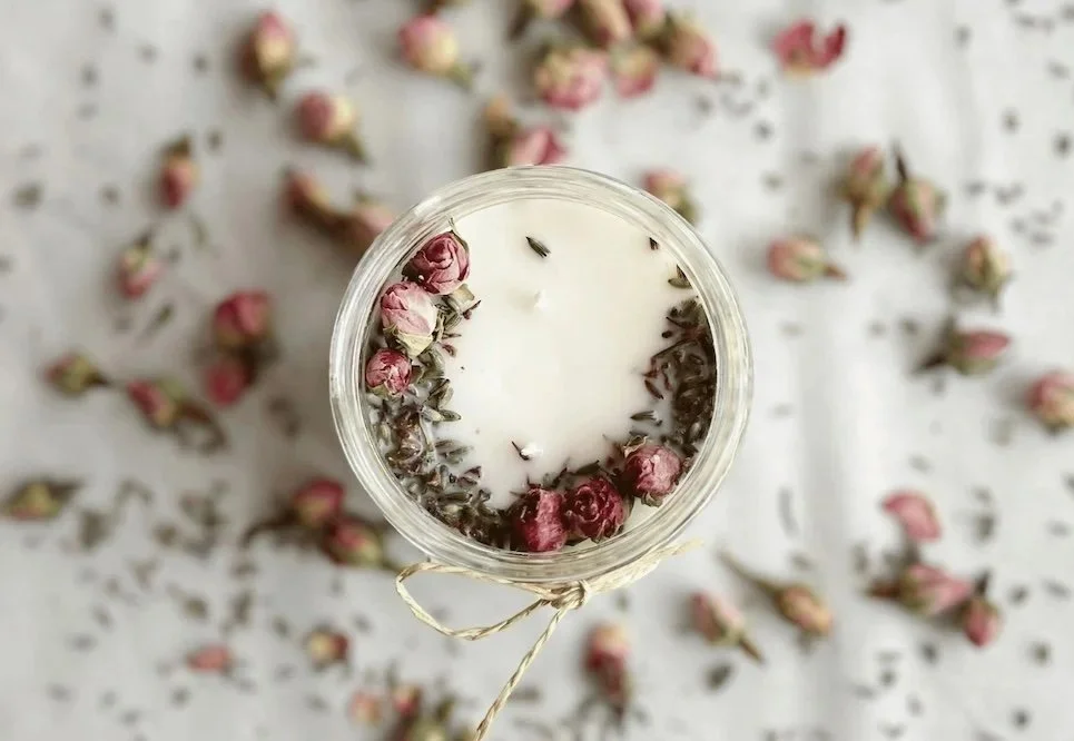 A white candle with dried rosebuds around the edge, placed in a glass jar, with more rosebuds scattered around on a white surface.