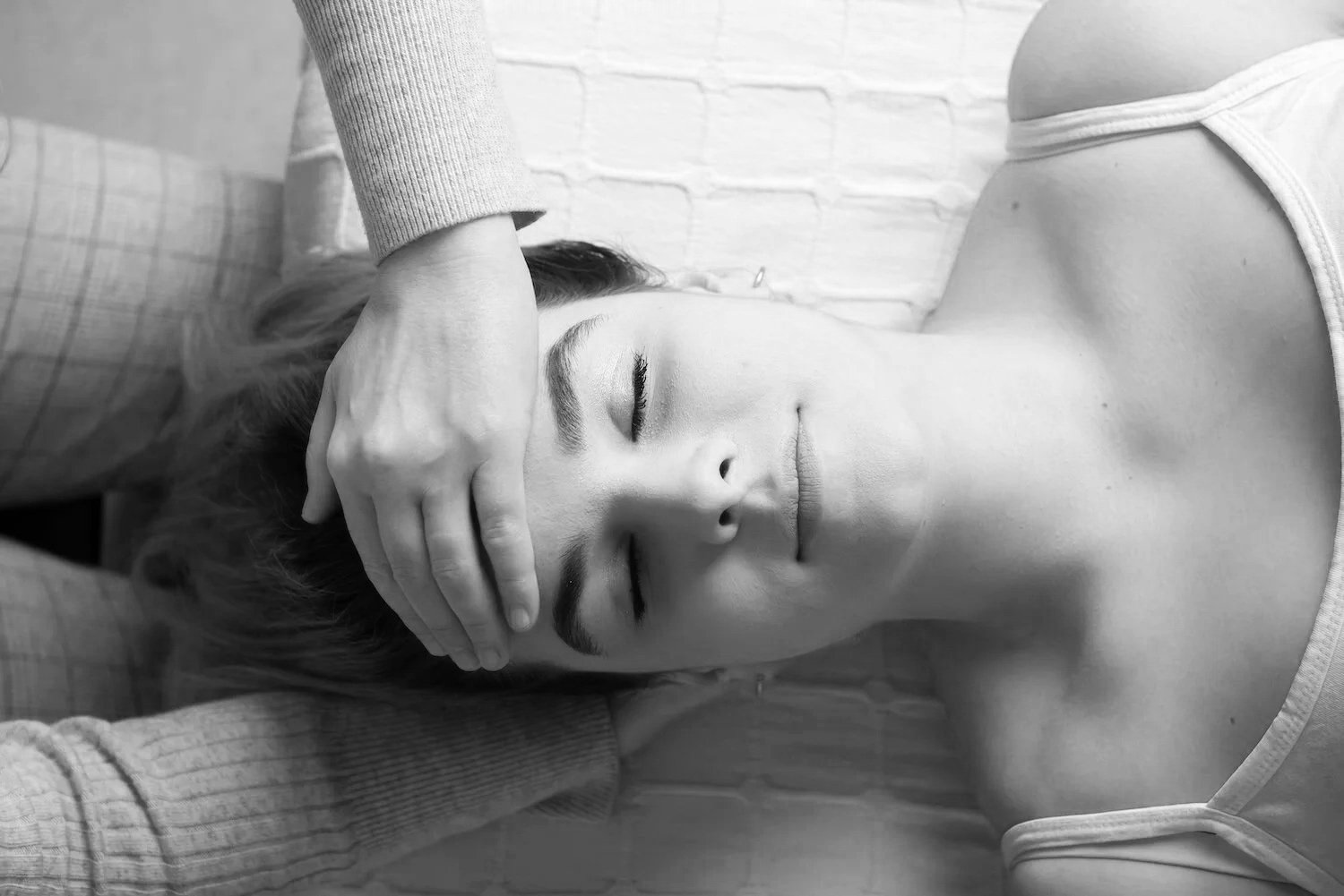 A person lying down with eyes closed, receiving a gentle craniosacral head treatment from someone with hands on their forehead.