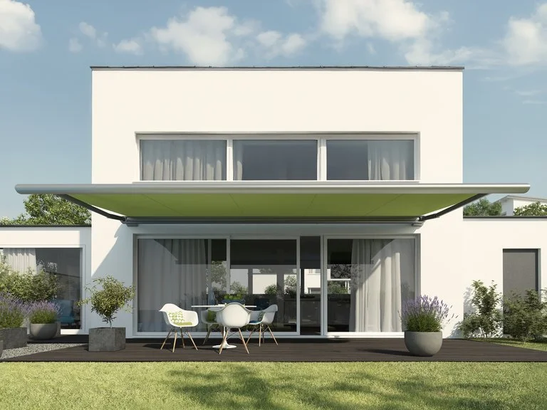 Modern two-story white house with large glass windows and sliding doors, an extended green awning shading the patio, and outdoor furniture on a wooden deck, surrounded by potted plants and a grassy lawn.