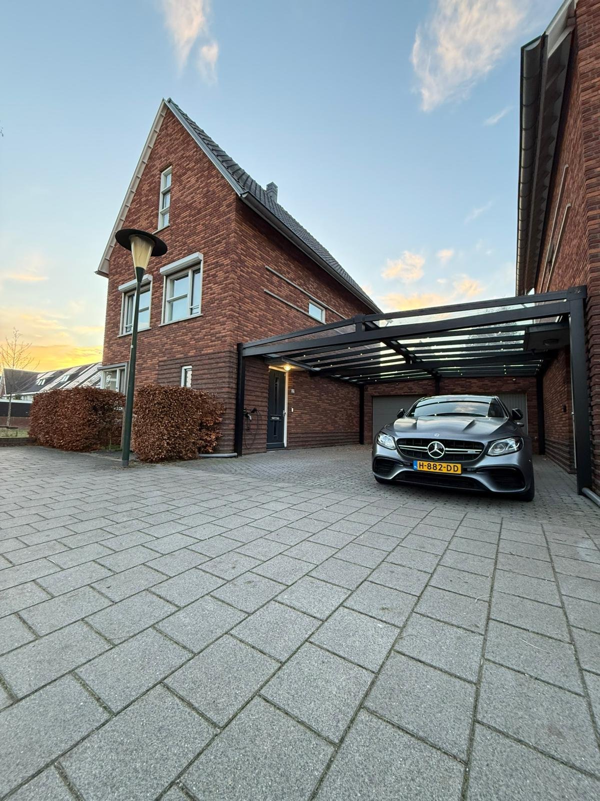 View of a modern brick house with a driveway, a black Mercedes-Benz parked under a carport, bushes, and a streetlamp at sunset.