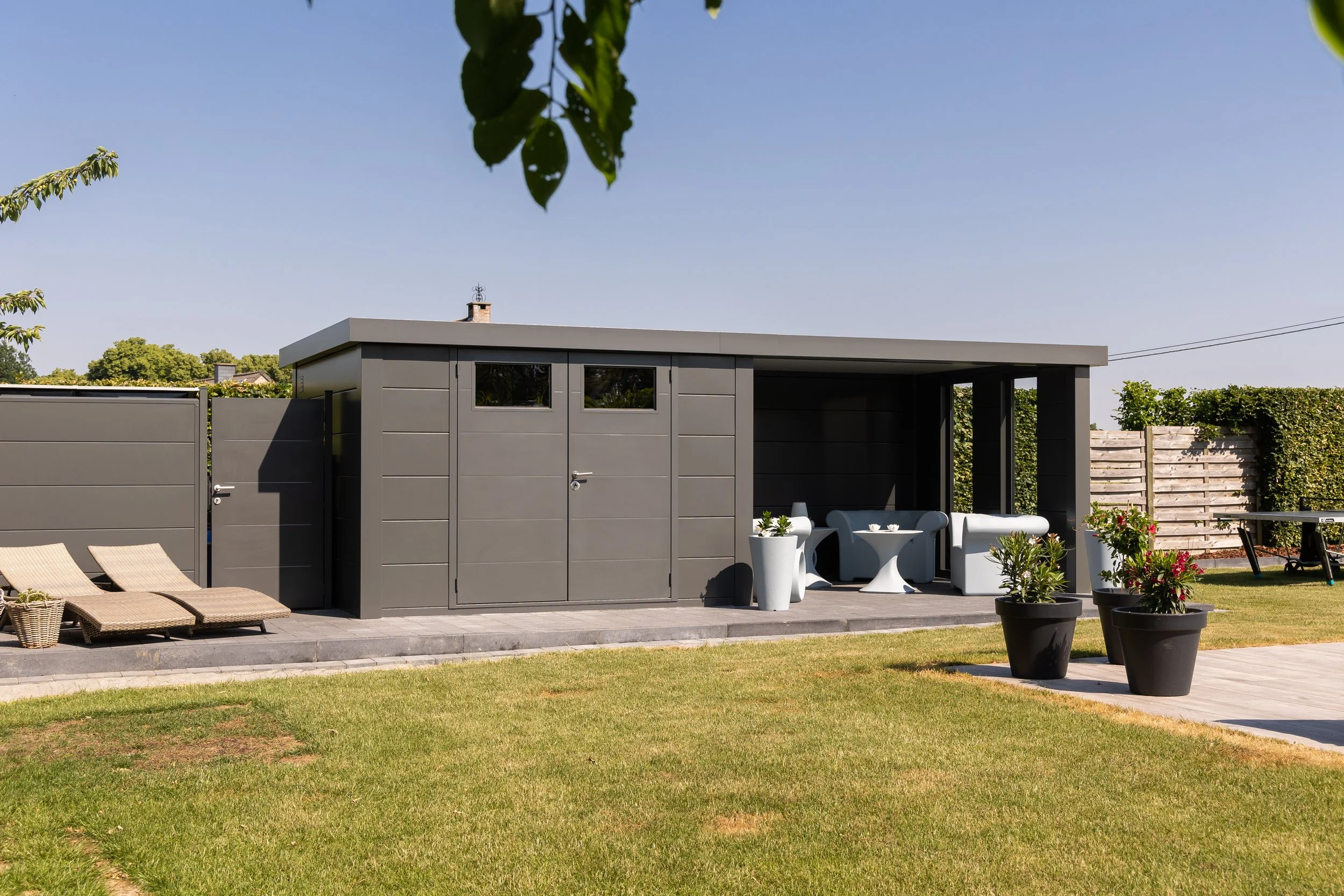 Grey outdoor shed with patio furniture and potted plants on a well-kept lawn under a clear blue sky.
