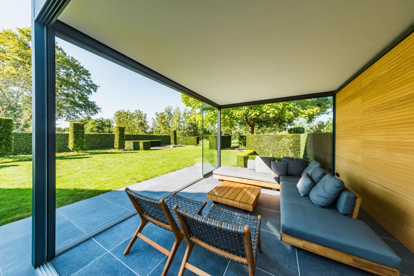 Modern glass-enclosed porch with outdoor furniture, including a gray sectional sofa with pillows, a wooden coffee table, and two woven chairs, overlooking a lush green yard with hedge landscaping.