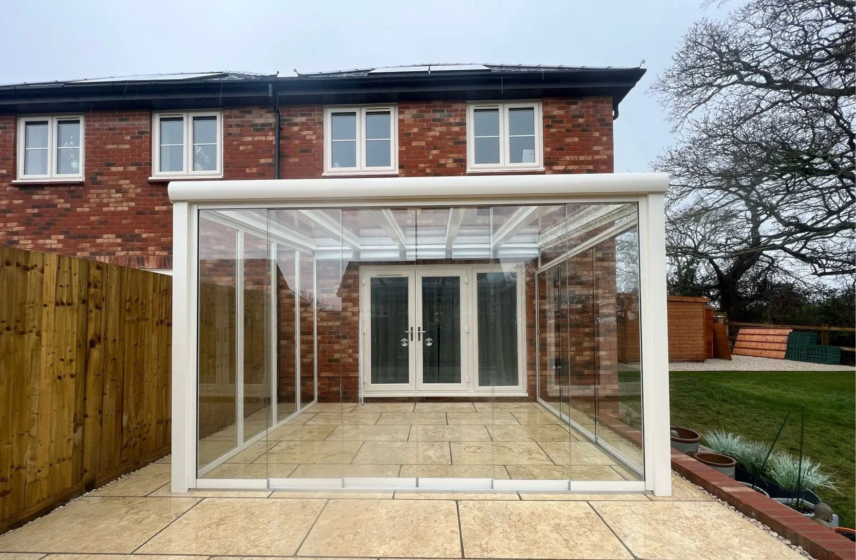 Backyard patio with a glass conservatory attached to a brick house, wooden fence on the left, trees and green lawn on the right.