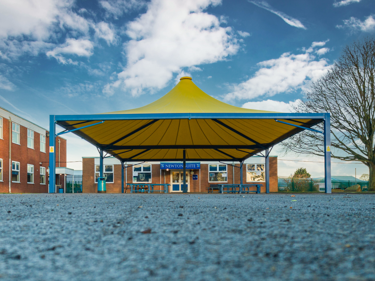 A yellow and blue canopy tent in front of a brick building with a sign that reads 'Newton Suite', outdoor tables and benches, and a clear sky with scattered clouds.