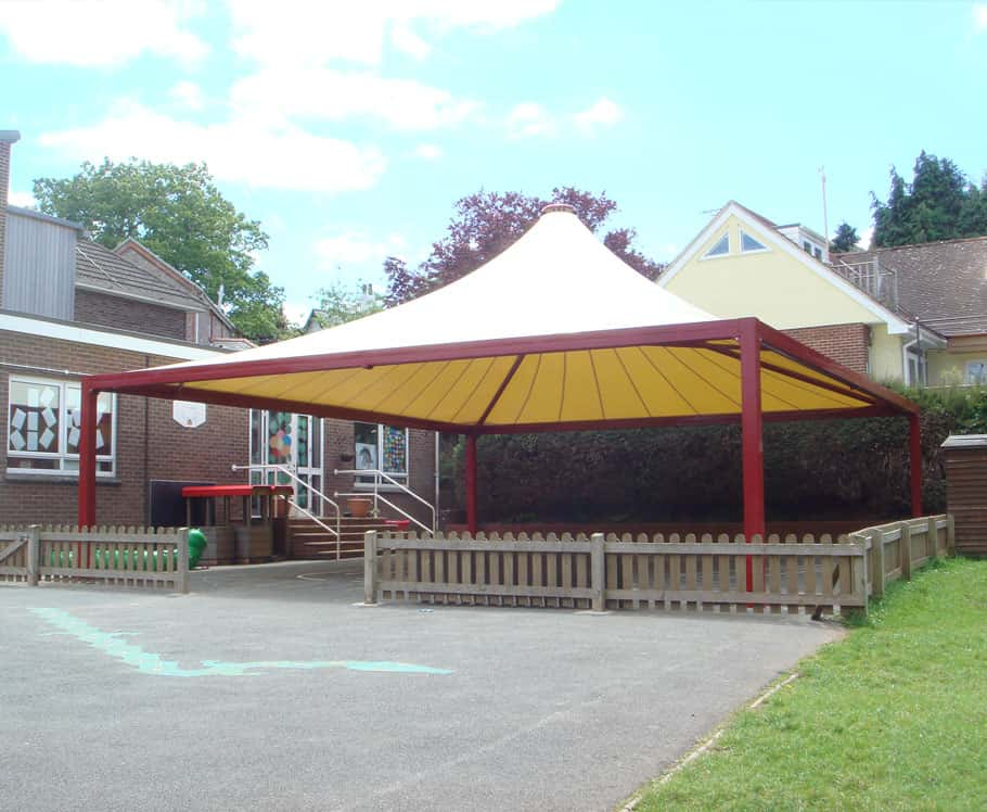 School playground with a large yellow and red canopy, a basketball hoop, and a fenced area for children to play.