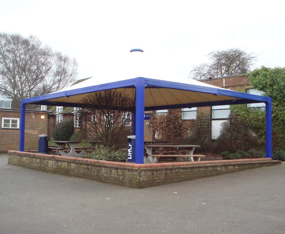 An outdoor shelter with a blue frame and beige canopy, covering picnic tables and benches, situated on a raised brick platform surrounded by plants and trees.