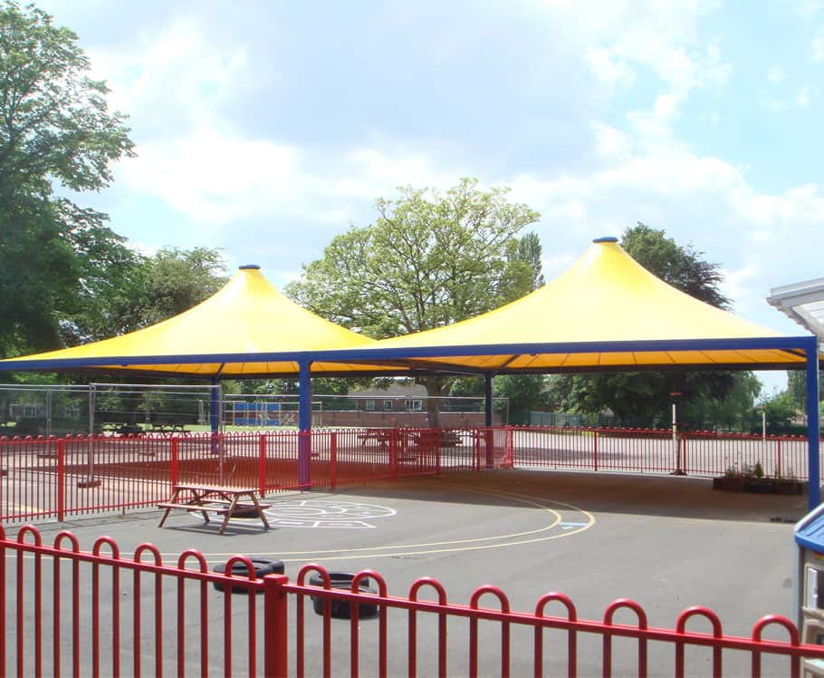 An outdoor playground with a yellow canopy structure, surrounded by red fencing, a picnic table, and trees in the background.