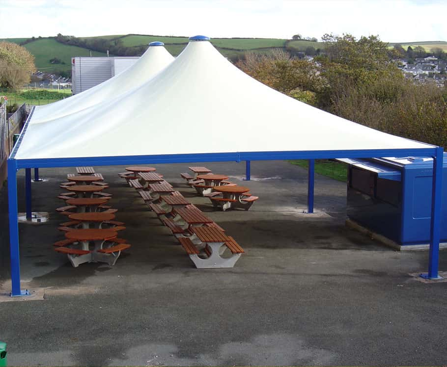 An outdoor seating area under a large white tent with blue supports, featuring wooden picnic tables, beside vending machines, on a paved surface with hills and trees in the background.