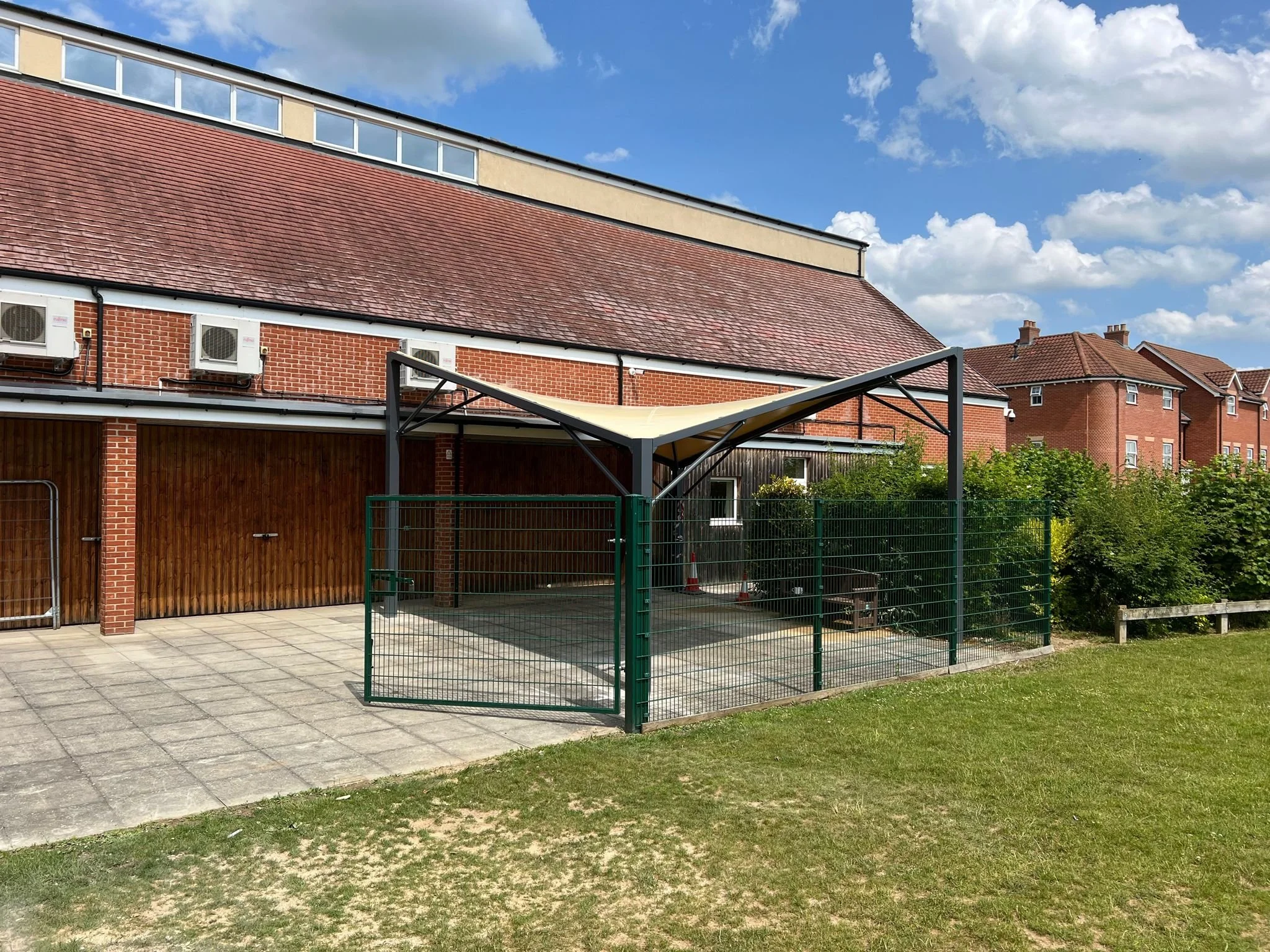 Outdoor area with a brick building, a covered shelter with a fabric roof, a metal fence, and a grassy lawn under a partly cloudy sky.