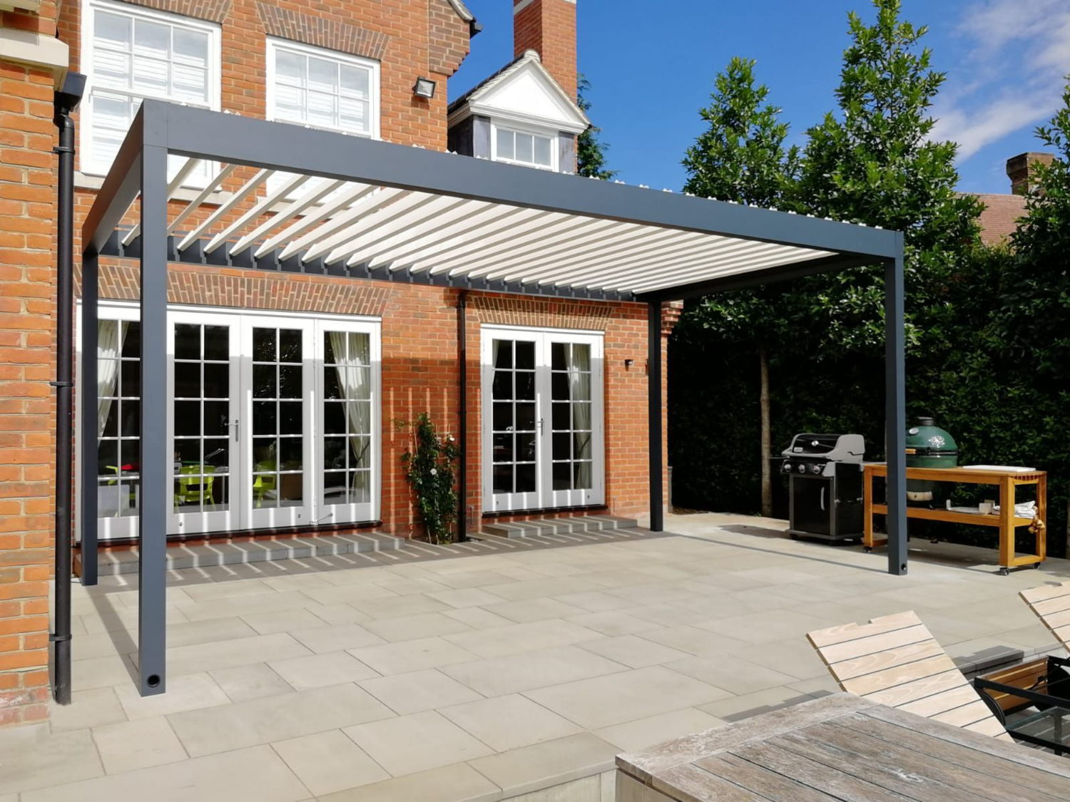 Backyard patio with a modern gray pergola, glass doors with white curtains, potted plants, a barbecue grill, a wooden table, and outdoor chairs, under a partly cloudy sky.