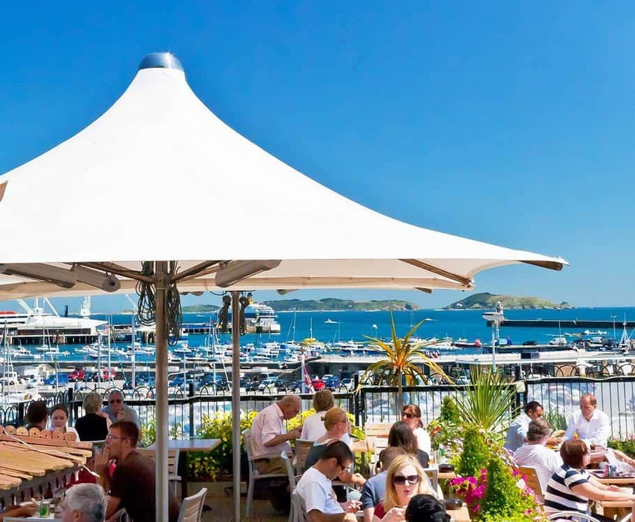 People dining outdoors at a waterfront restaurant with a marina full of boats in the background, under a large white canopy in a sunny setting.