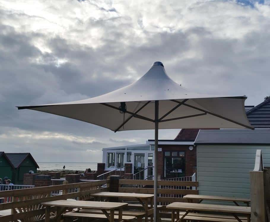 Outdoor patio with wooden picnic tables and a large white patio umbrella, overlooking a beach and ocean under cloudy skies.