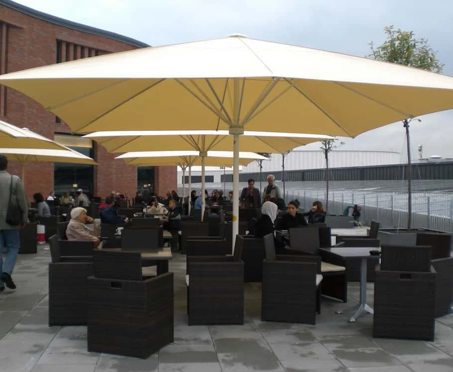 Outdoor seating area with black wicker chairs and dark tables under large beige umbrellas at a busy café or restaurant.