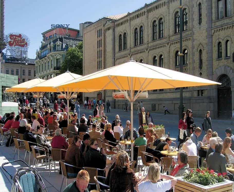 People dining at outdoor tables under umbrellas on a city street with historic buildings and commercial signs in the background.