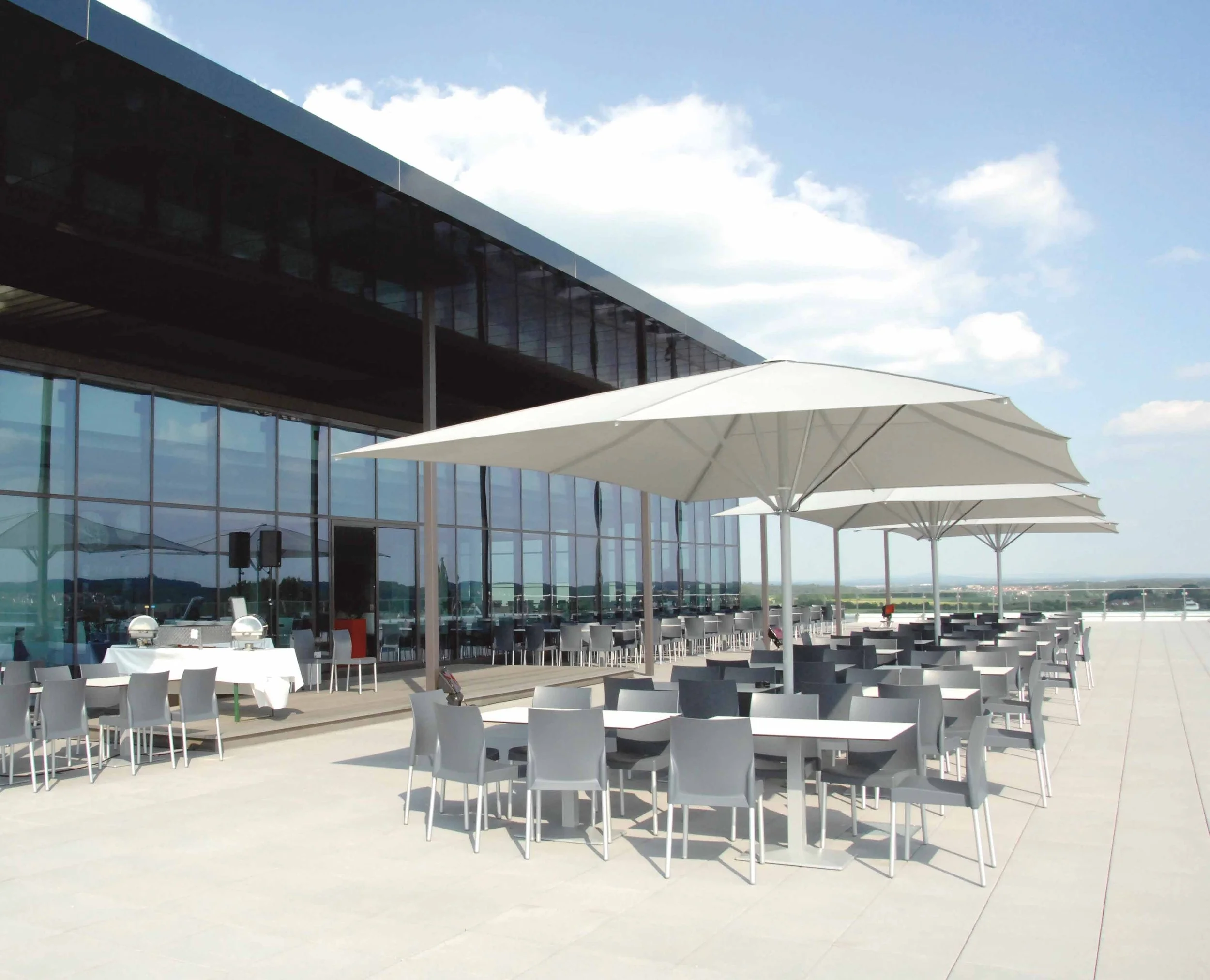 Outdoor rooftop dining area with white tables and gray chairs under large white umbrellas, adjacent to a modern glass building, with a clear sky and distant landscape in the background.