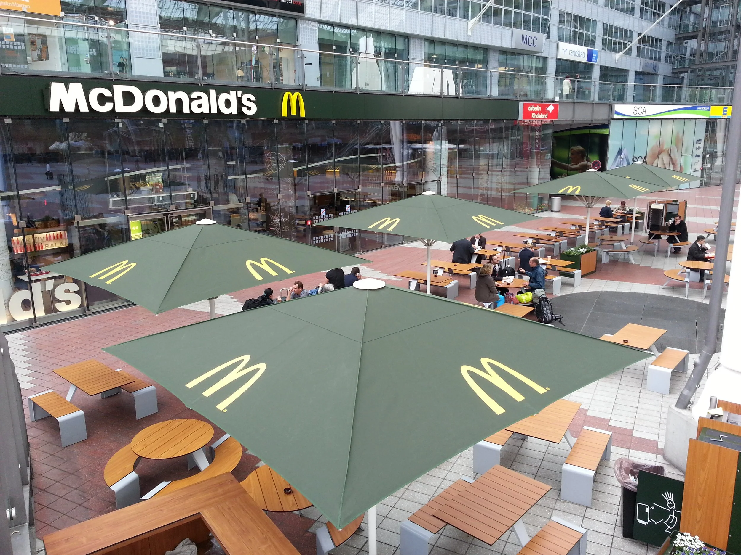 Outdoor seating area of a McDonald's restaurant with tables, benches, large green umbrellas with yellow McDonald's logos, and people dining in and waiting outside, in front of a glass building with multiple signs.