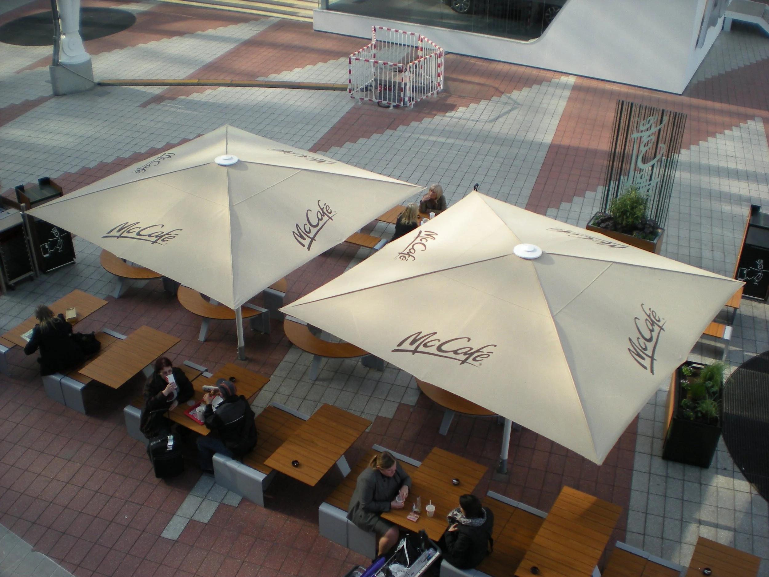 Outdoor seating area with McDonald's McCafé umbrellas, wooden tables, and customers eating and drinking.