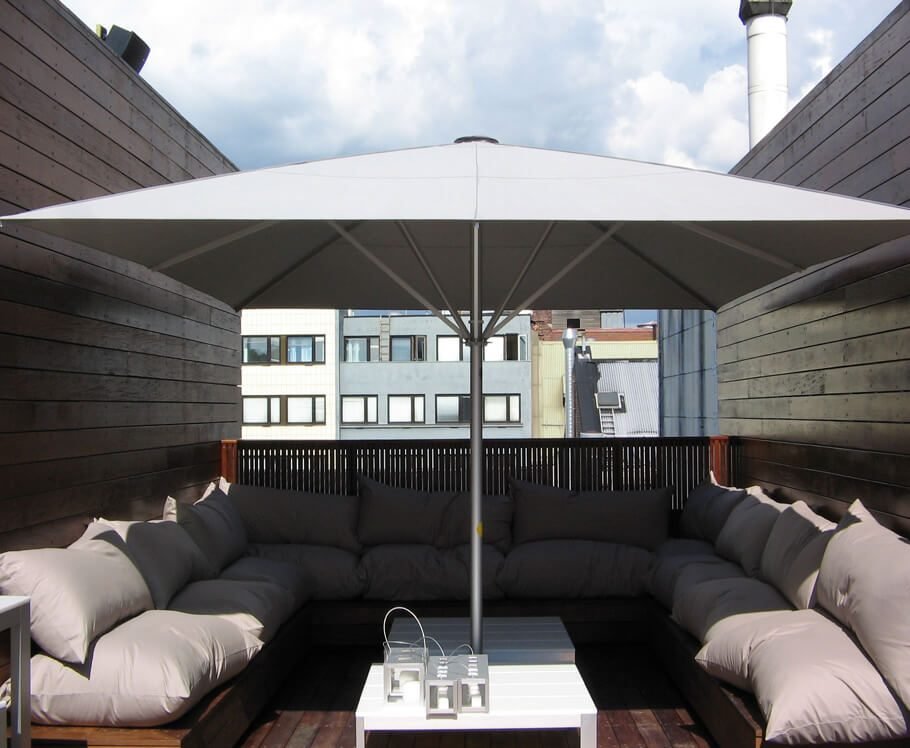 Outdoor rooftop patio with a large white umbrella, sectional sofa with cushions, and a white coffee table holding lanterns, surrounded by wooden walls and urban buildings in the background.