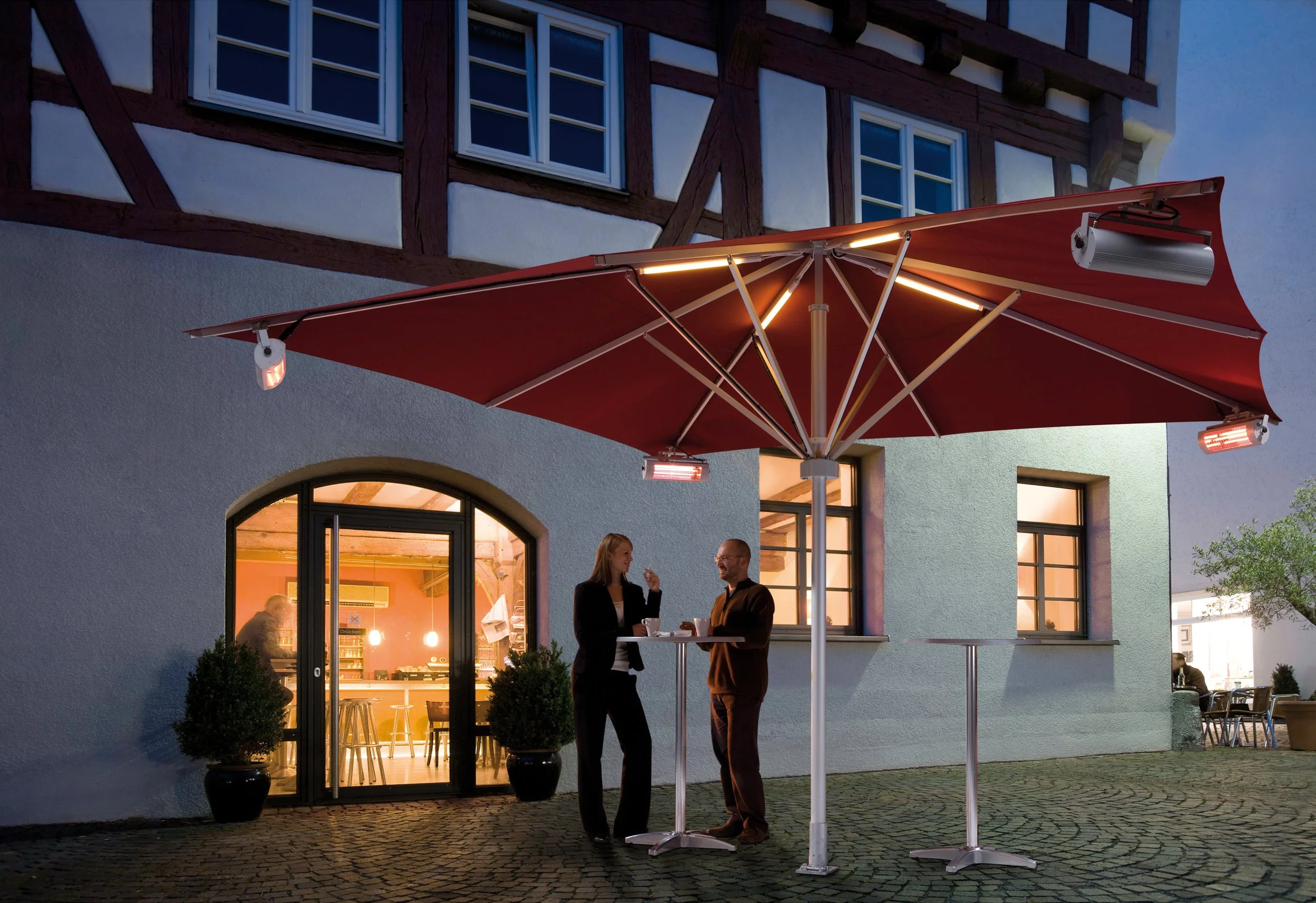 Two people, a woman and a man, standing and talking under a red patio umbrella outside a restaurant or cafe at night, with warm indoor lighting and potted plants visible through the windows.