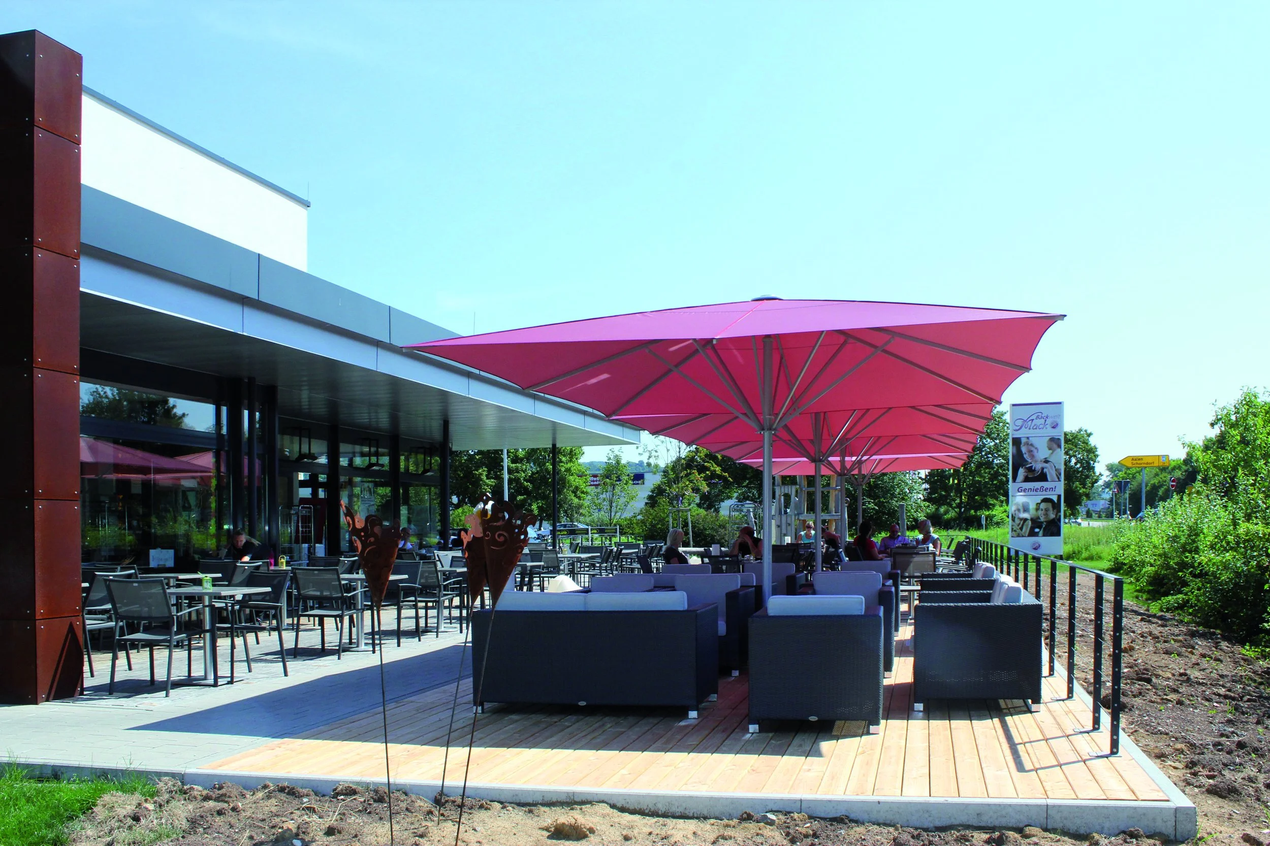 Outdoor seating area of a cafe or restaurant with black chairs and tables, large pink umbrellas, and a few people sitting, next to a modern building with glass windows, on a sunny day.