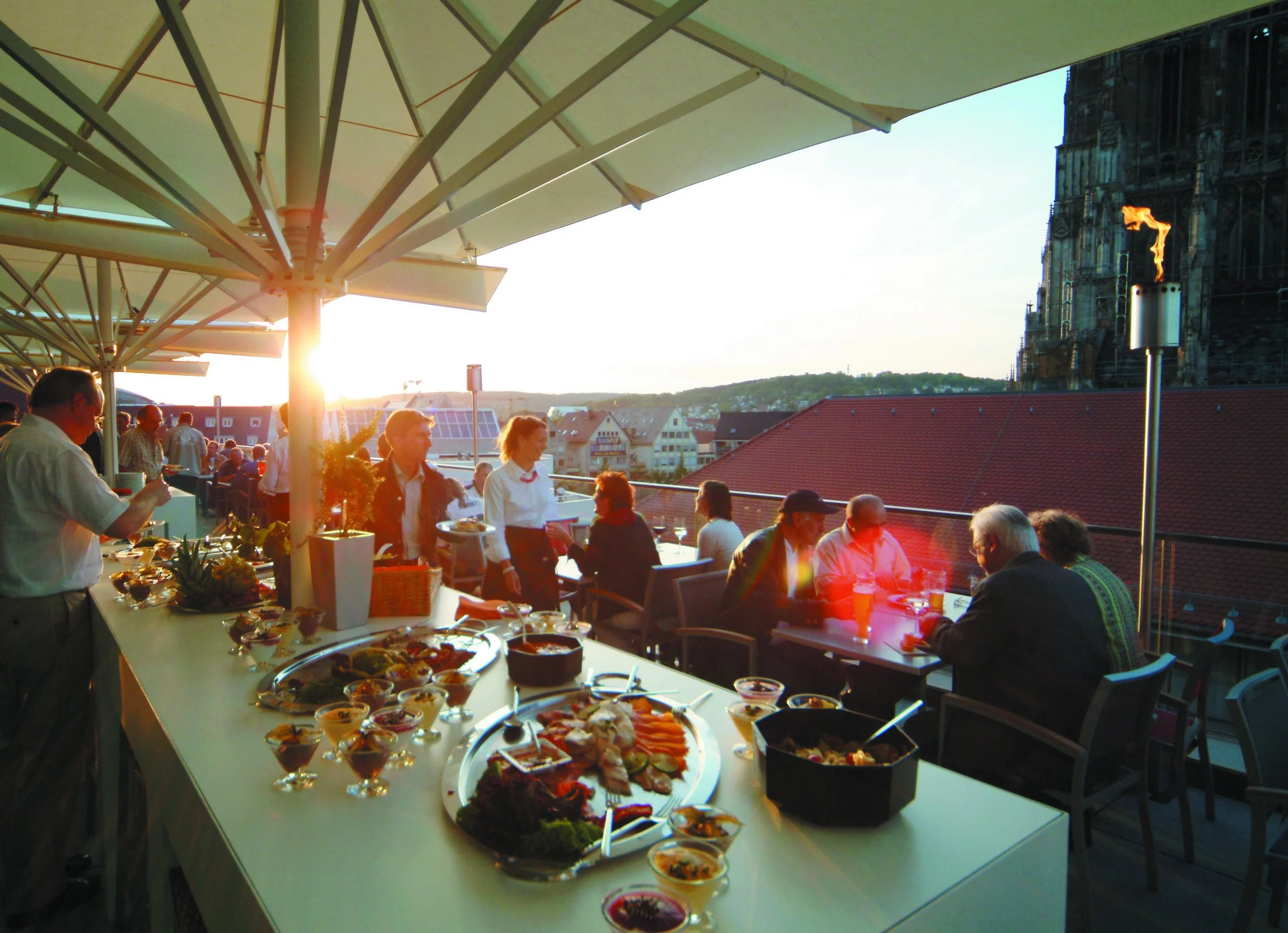People dining on a rooftop terrace during sunset, with a variety of food on a large table in the foreground and a historic church tower in the background.