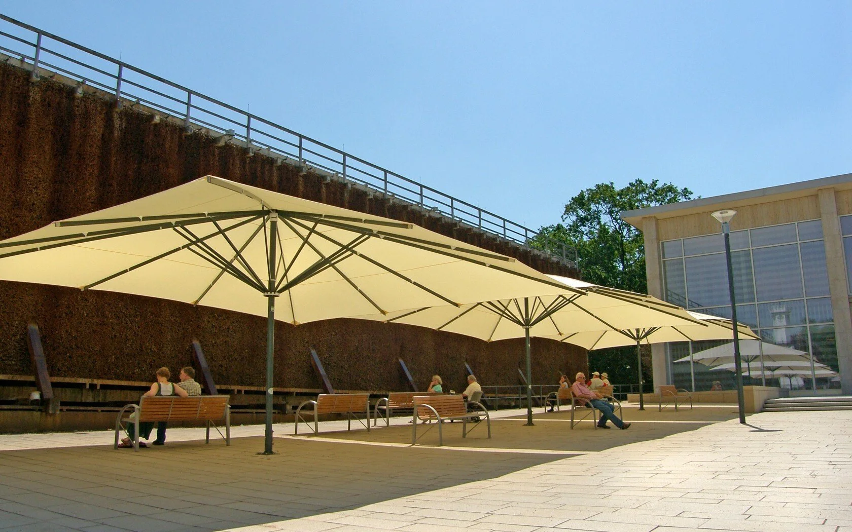 A modern outdoor patio area with large beige umbrellas, benches, several people sitting and talking, surrounding buildings and a clear blue sky.