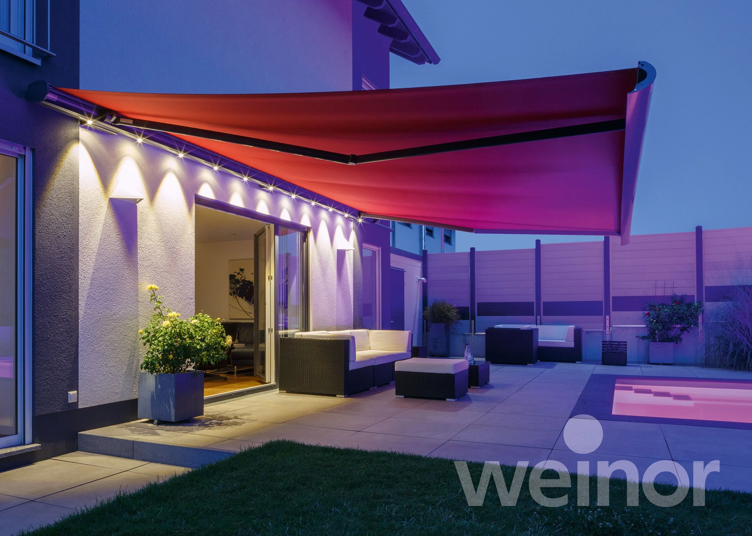 Modern backyard patio with outdoor seating, potted plants, sliding glass door, illuminated wall lights, and a retractable awning at dusk.
