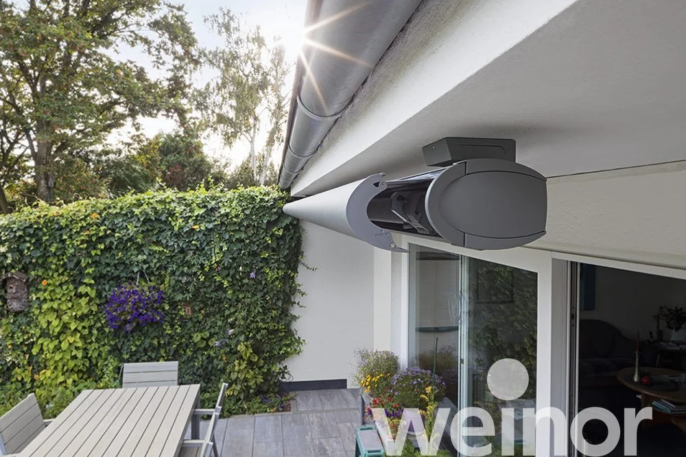 Close-up of a retractable outdoor awning rolled out above a patio with garden furniture and potted plants, surrounded by a green hedge and trees, in sunlight.