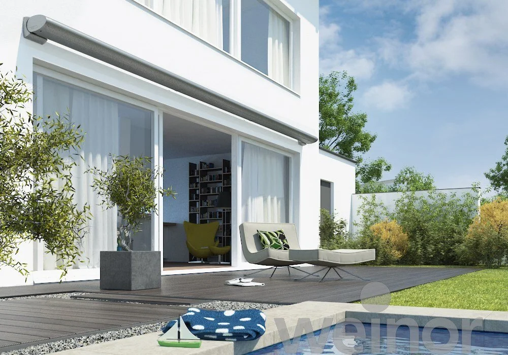Modern white house with large glass sliding doors open to a patio with outdoor lounge chair, potted tree, and a swimming pool surrounded by greenery and blue sky.