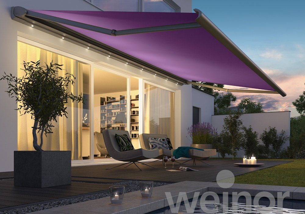 Modern outdoor patio with two lounge chairs, a potted tree, a purple retractable awning, and a view of a house with interior lighting, a bookshelf, and sliding glass doors at sunset.