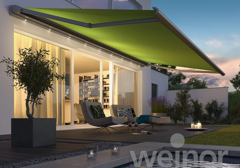 Modern patio with sliding glass doors, outdoor lounge chairs, a potted tree, candles, and a green retractable awning at sunset.