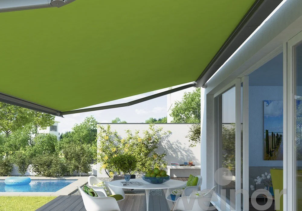 Outdoor patio with white table and green cushion chairs, fruit bowl, pool in the background, and a green awning providing shade.