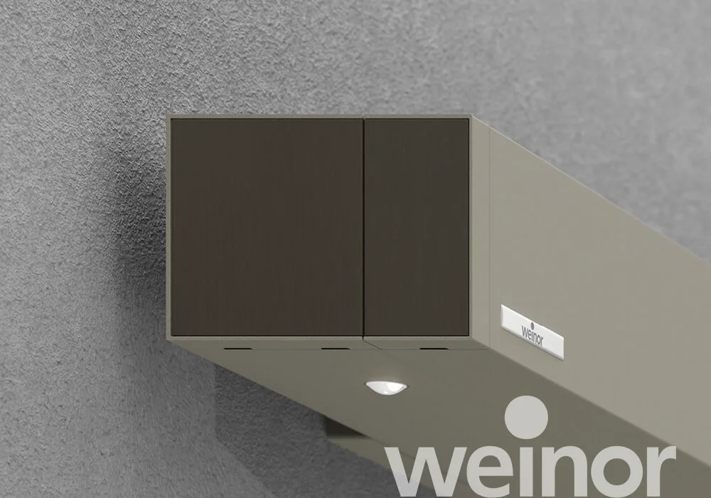 Close-up of a gray ceiling-mounted air vent with dark brown louvers and a nearby illuminated smoke detector on a gray ceiling with a textured surface. The vent has a small label with the brand name 'Weinor' and the logo is partly visible in the botto