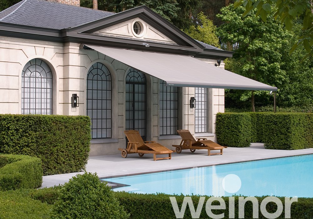 Residential house with large arched windows, a pool with a surrounding deck, two wooden lounge chairs, a gray retractable awning, and green manicured bushes and trees.