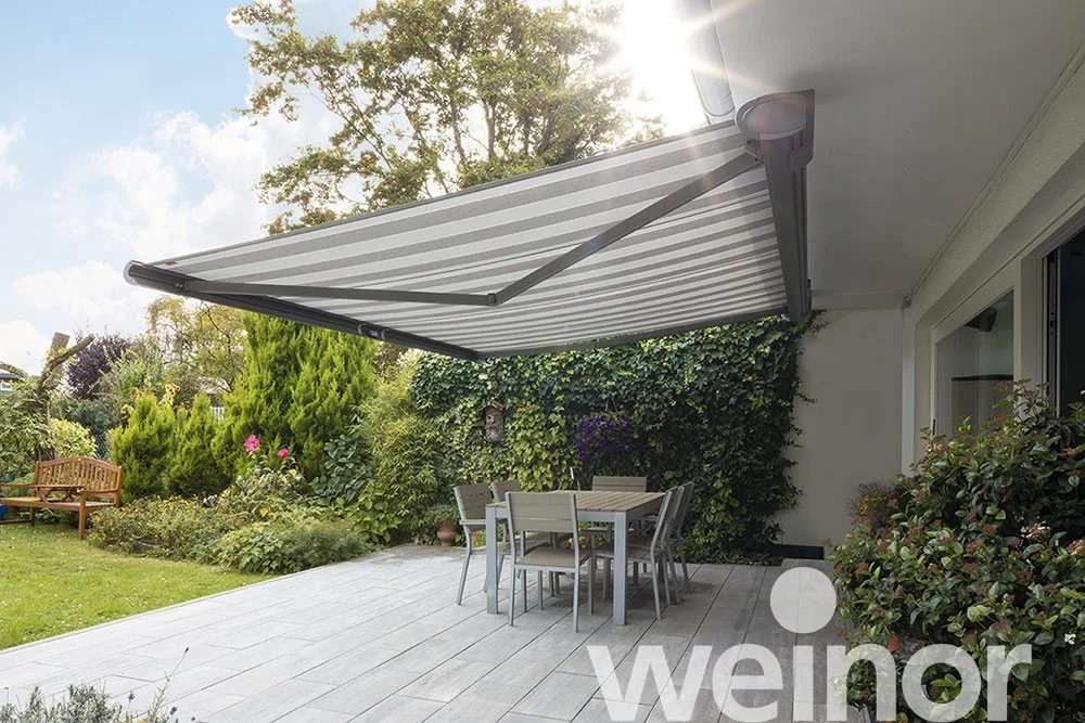 Outdoor patio with a retractable striped awning, a table with six chairs, a garden with lush trees and plants, and a wooden bench in a backyard.