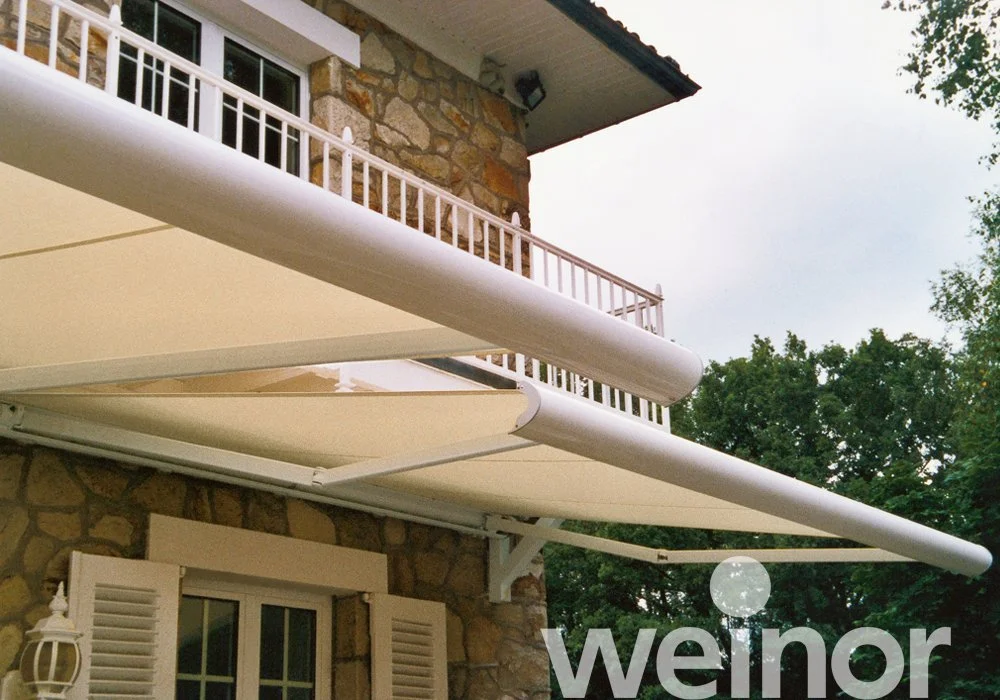 Exterior view of a stone house with a balcony, white railings, window shutters, and adjustable beige awnings extended outside.