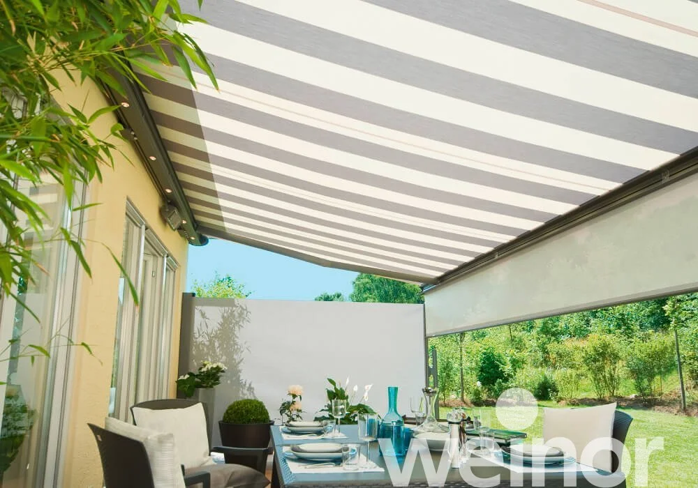 Outdoor patio with dining table set for a meal, surrounded by greenery and shaded by a striped retractable awning.