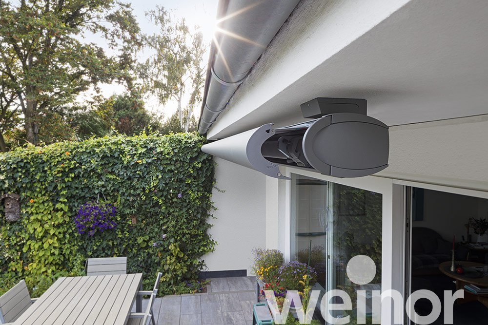 Close-up of an outdoor wind or security sensor mounted on the exterior wall of a house with a sliding door, overlooking a patio with outdoor furniture and lush green hedge.