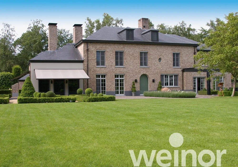 Large brick house with multiple windows, a green front door, a well-manicured lawn, and trimmed hedges, set against a backdrop of trees.