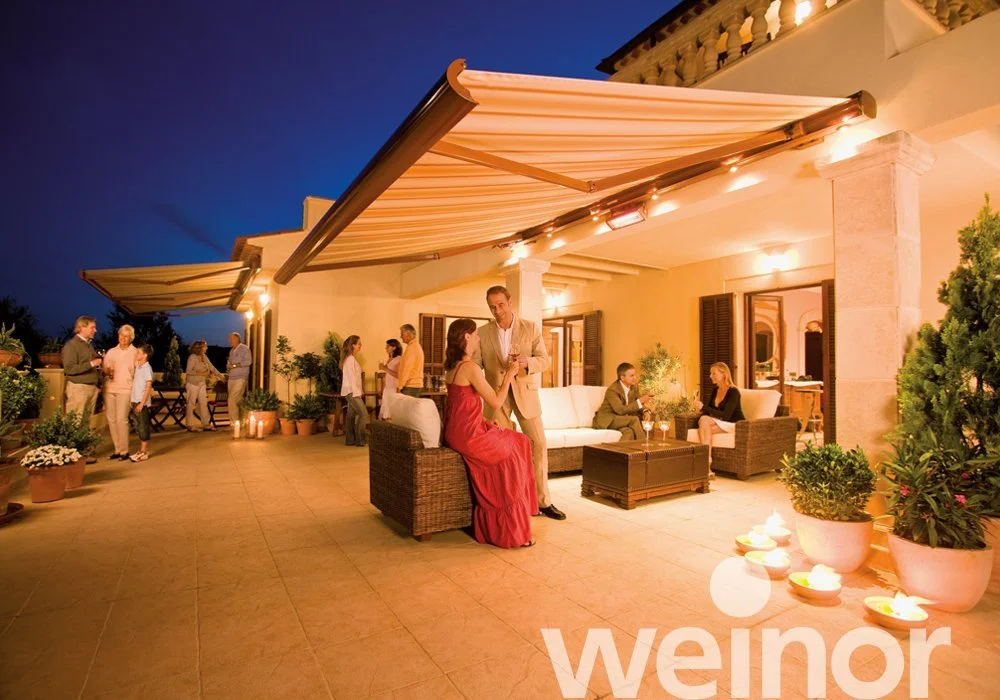 An outdoor evening gathering on a furnished terrace with people socializing, potted plants, string lights, and a retractable awning.