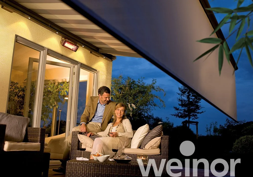 A man and woman enjoying drinks and conversation on a balcony or patio at dusk, with outdoor furniture, potted plants, and a view of trees.