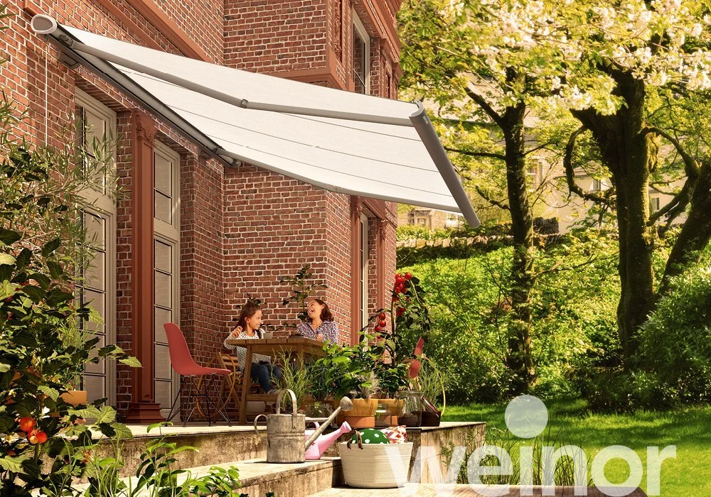 Two children sitting at a patio table on a backyard terrace with a white retractable awning, surrounded by lush green trees and plants, enjoying a sunny day.