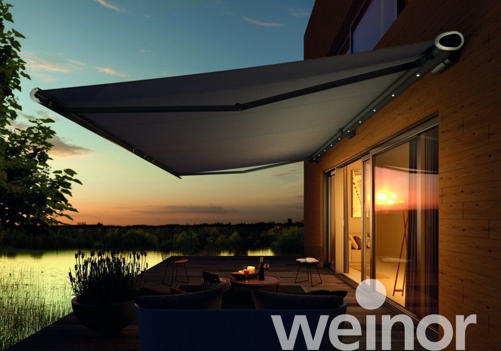 Sunset view from a balcony with an awning, outdoor seating, and a lake in the background.