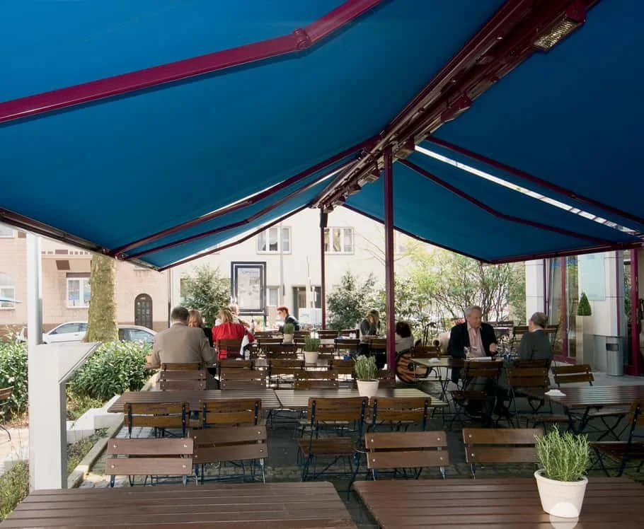 An outdoor seating area of a restaurant with wooden tables and chairs, some occupied by people, covered by a large blue patio umbrella, with trees and buildings in the background.