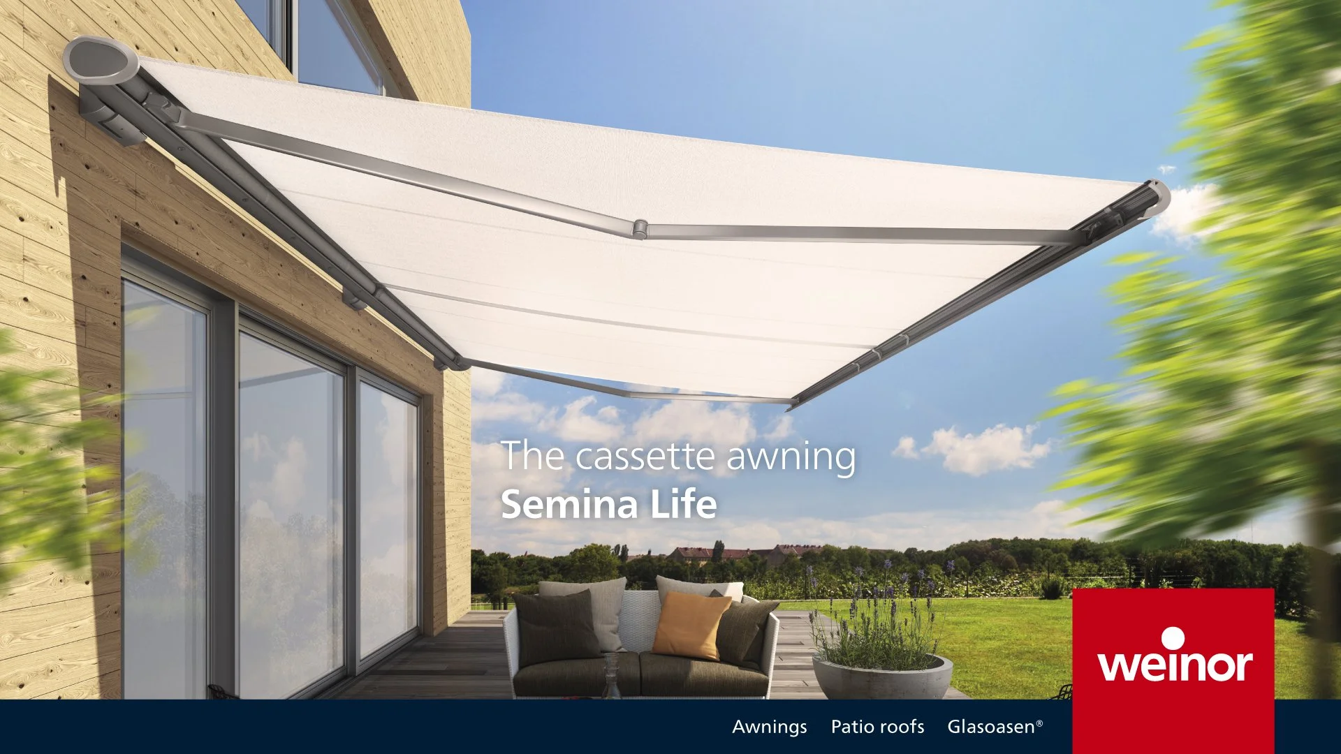 A white cassette-style retractable awning installed on a wooden house exterior over a patio with a sofa and a potted plant, with a grassy landscape and blue sky in the background.