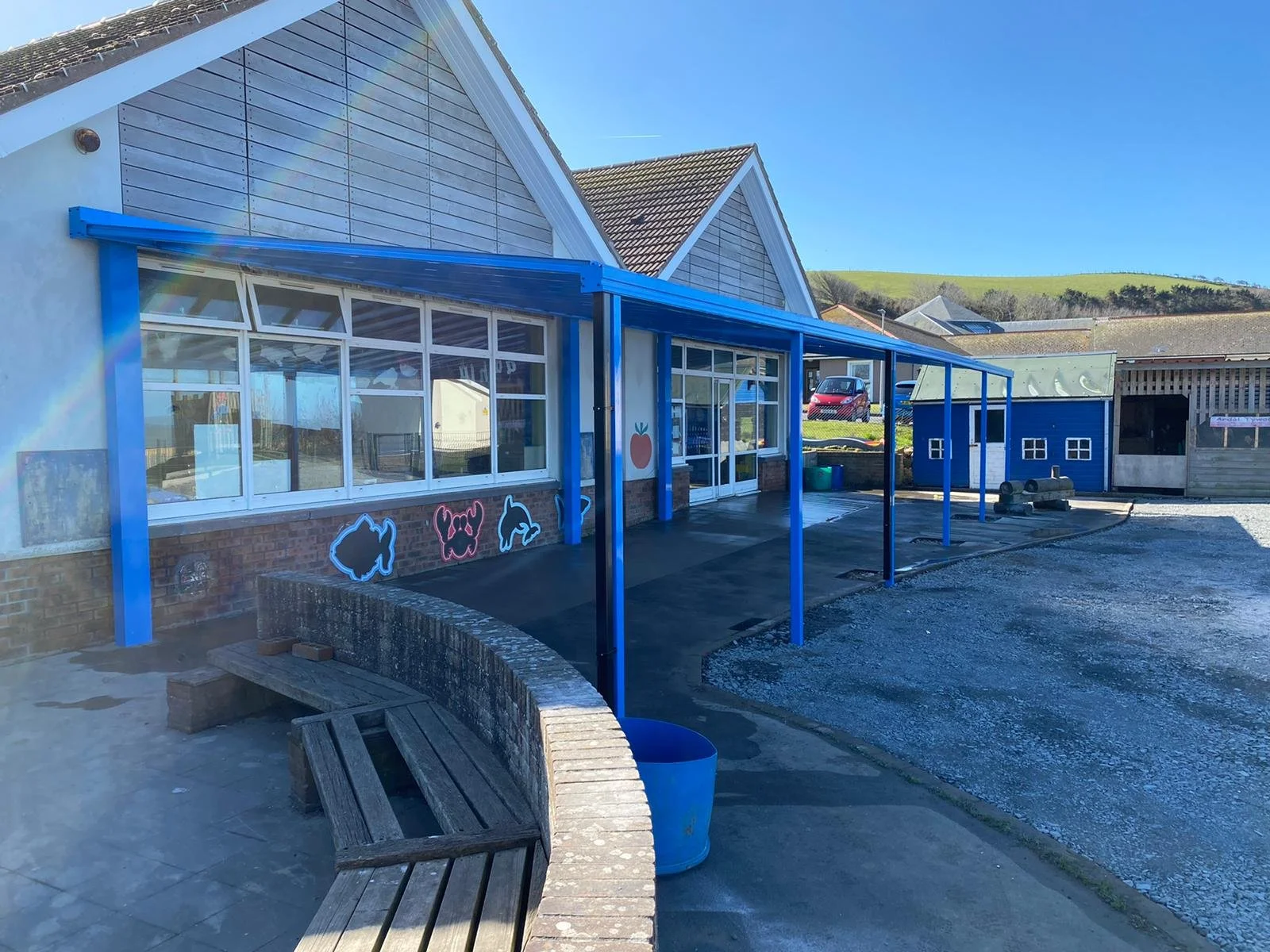 Outside of a building with a blue canopy and colorful animal depictions on the wall, a wooden bench, and a blue bucket in front. The building has large windows, a sloped roof, and a seaside or rural background with hills.
