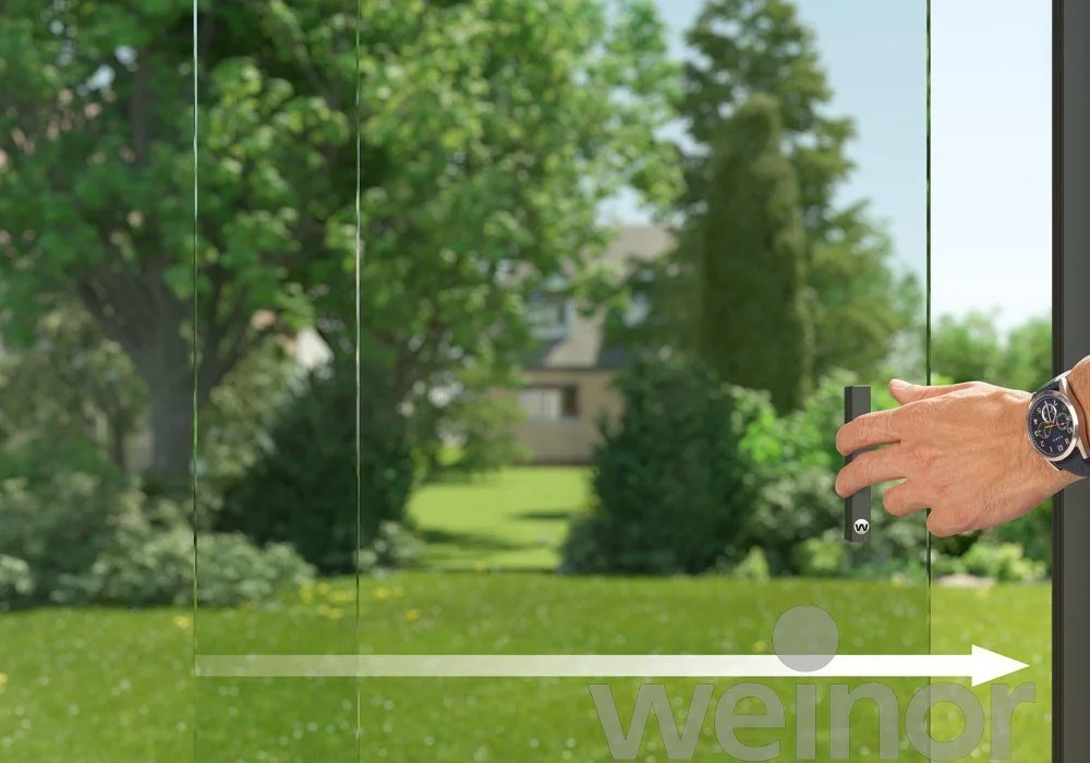 Person adjusting a glass door with a black handle, showing a green lawn, trees, and a house in the background outside.