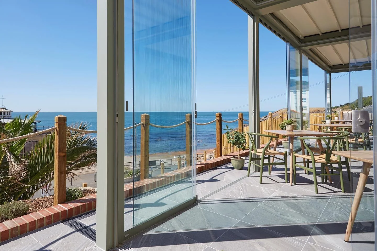 Indoor seating area with round tables and chairs overlooking a balcony with a view of the ocean and blue sky.